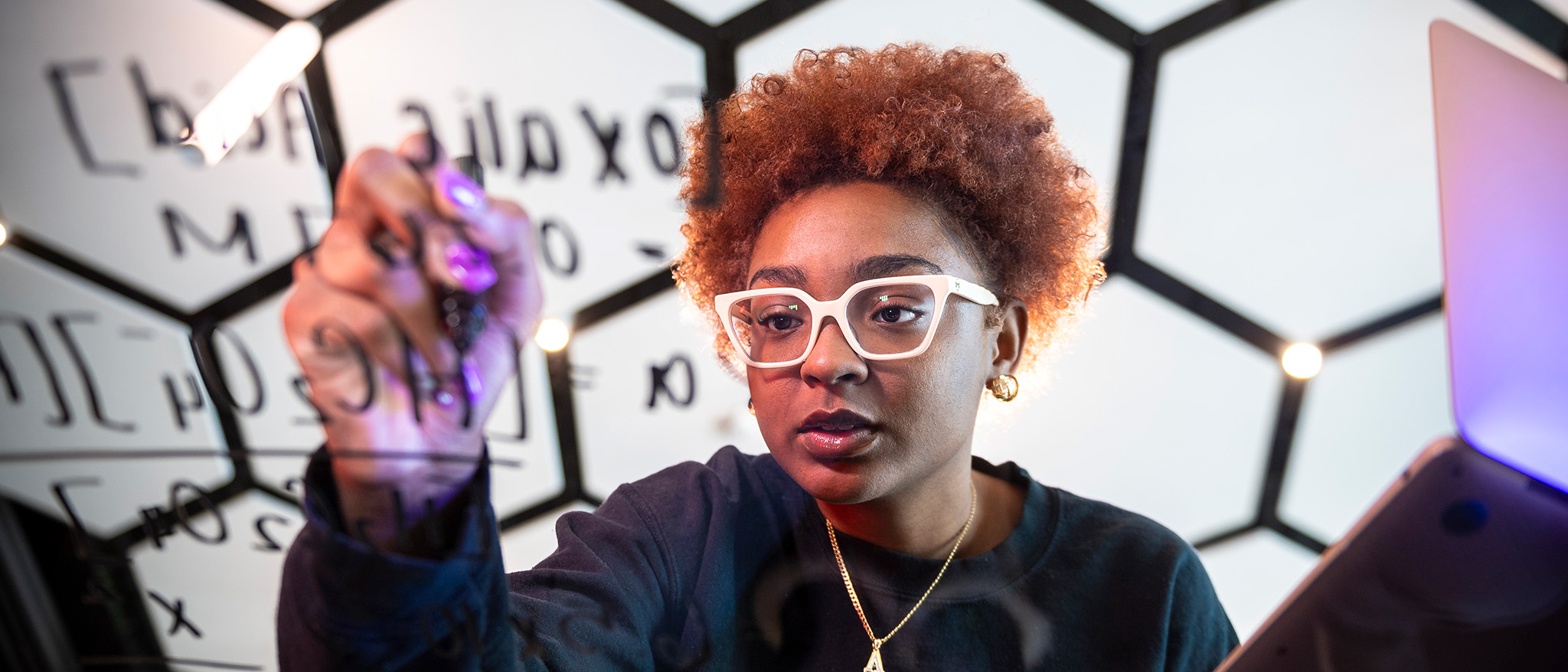 Students writes a math equation on a clear surface in front of a black and white honeycomb background