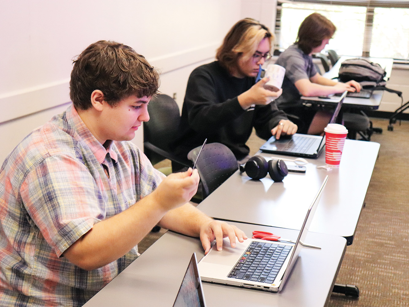 Three students sit at a long table in front of laptops