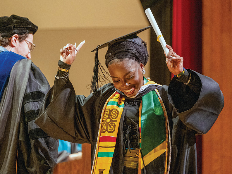 A woman in graduation regalia holds up her diploma and her pointer finger in celebration