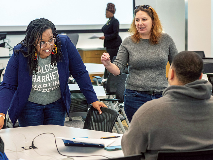 Two professors stand at a classroom table, speaking to a student who is sitting down