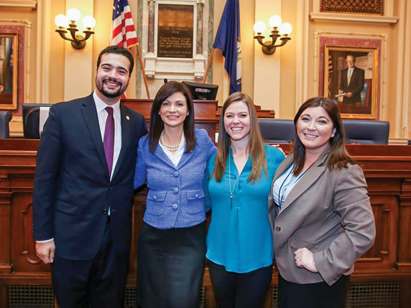 A man and three women dressed in business attire stand inside the Virginia State Capitol